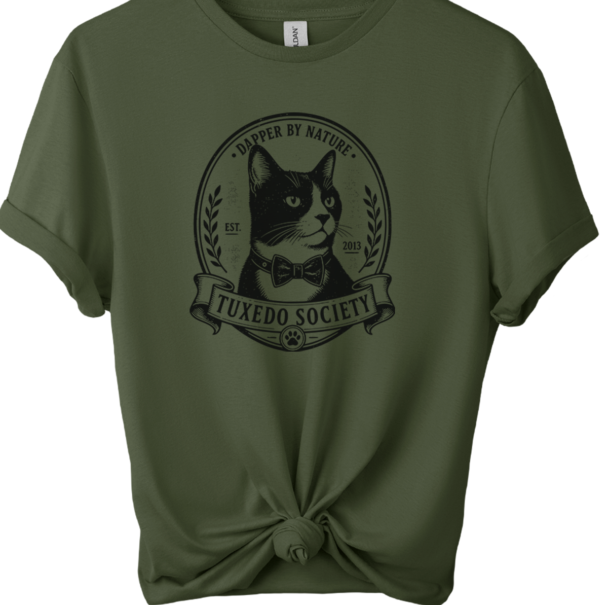 An military green short-sleeve t-shirt on a wooden hanger, styled with a knot at the bottom hem. The shirt features a black graphic print of a cat wearing a bow tie inside an oval frame with the text 'TUXEDO SOCIETY' and dapper BY NATURE'.