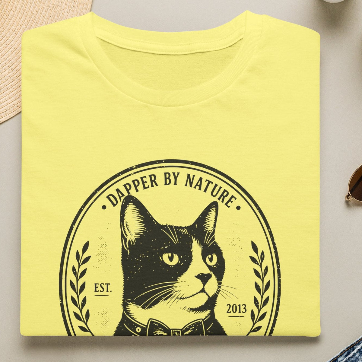 A folded cornsilk  T-shirt is displayed in a styled flat lay on a light beige surface. The shirt features a black graphic of a cat inside a circular emblem with the text 'DAPPER BY NATURE' and 'EST.