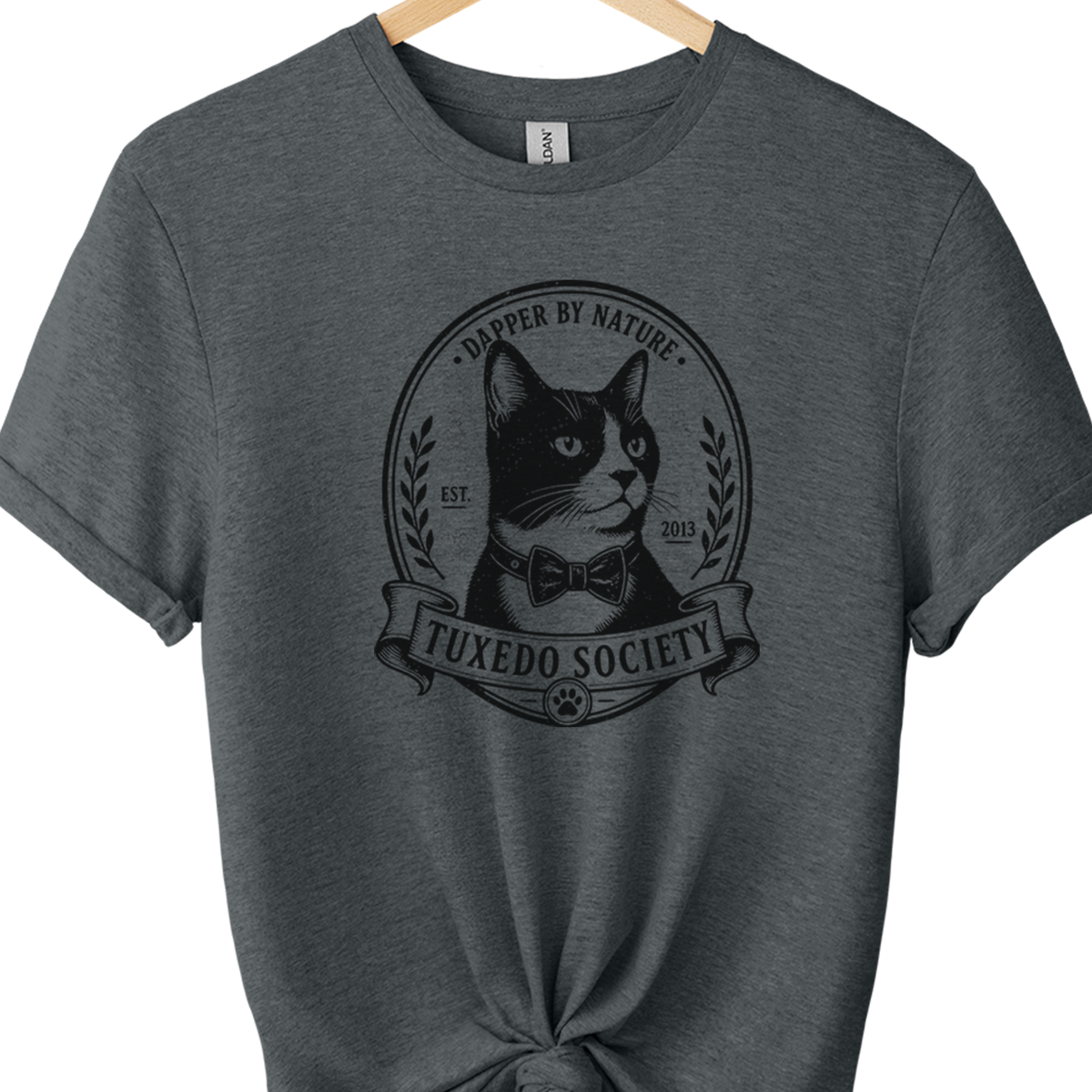 A dark heather gray t-shirt hanging on a wooden hanger against a white background, styled with a knot at the bottom hem and featuring a black graphic of a cat in a bow tie with the text 'TUXEDO SOCIETY' and 'A DOpper BY NAture'.