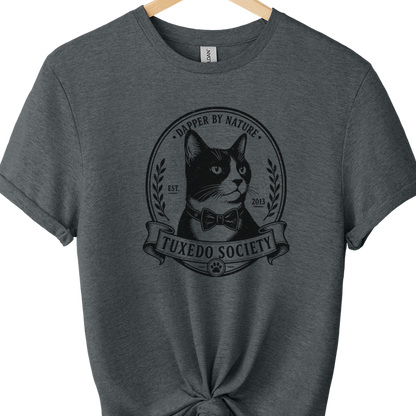 A dark heather gray t-shirt hanging on a wooden hanger against a white background, styled with a knot at the bottom hem and featuring a black graphic of a cat in a bow tie with the text 'TUXEDO SOCIETY' and 'A DOpper BY NAture'.