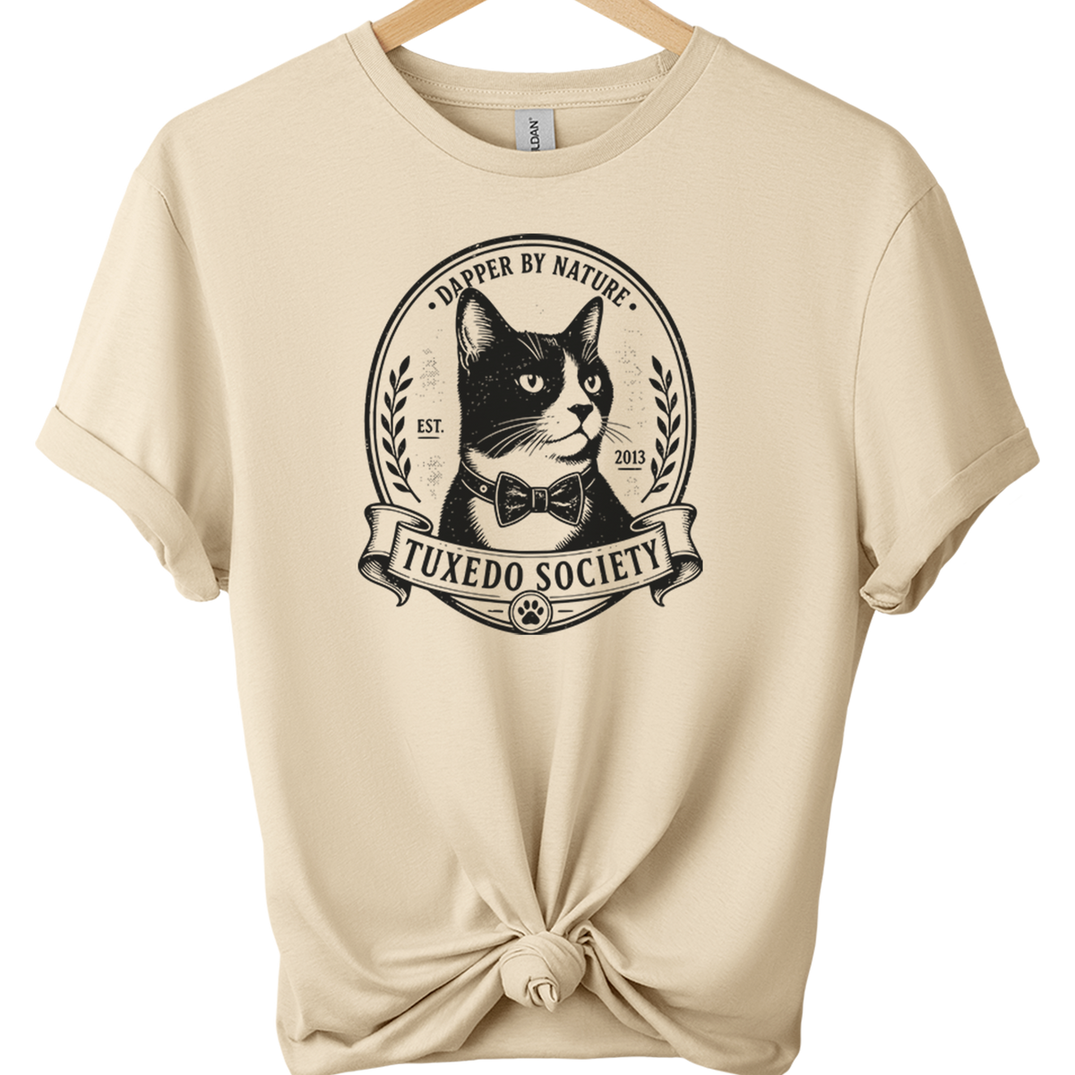A sand folded sleeve t-shirt on a wooden hanger, featuring a black graphic of a cat wearing a bow tie inside an oval emblem with the text 'TUXEDO SOCIETY' and 'TRAPPER BY NATURE'. The bottom of the shirt is tied in a knot.