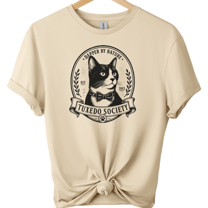 A sand folded sleeve t-shirt on a wooden hanger, featuring a black graphic of a cat wearing a bow tie inside an oval emblem with the text 'TUXEDO SOCIETY' and 'TRAPPER BY NATURE'. The bottom of the shirt is tied in a knot.