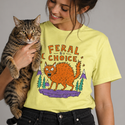 A woman holding a tabby cat wears a cornsilk graphic T-shirt with the text 'FERAL BY CHOICE' and an orange cat illustration, paired with black jeans, against a plain gray studio background.