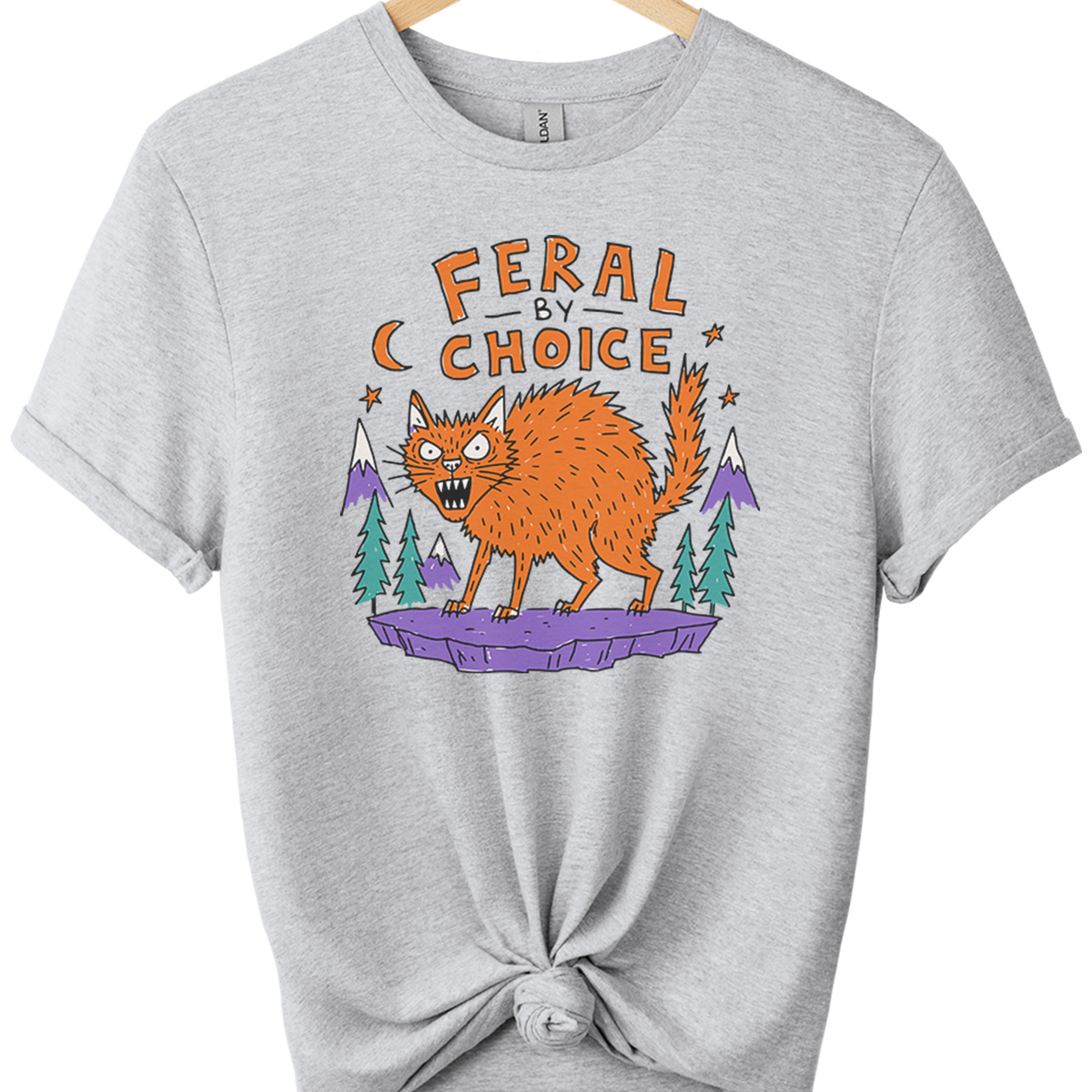 A light sport gray t-shirt hanging on a wooden hanger, styled with a knot at the bottom hem. The shirt features a graphic print of an orange feral cat on a purple platform with pine trees and the text 'FERAL BY CHOICE'.neilikha