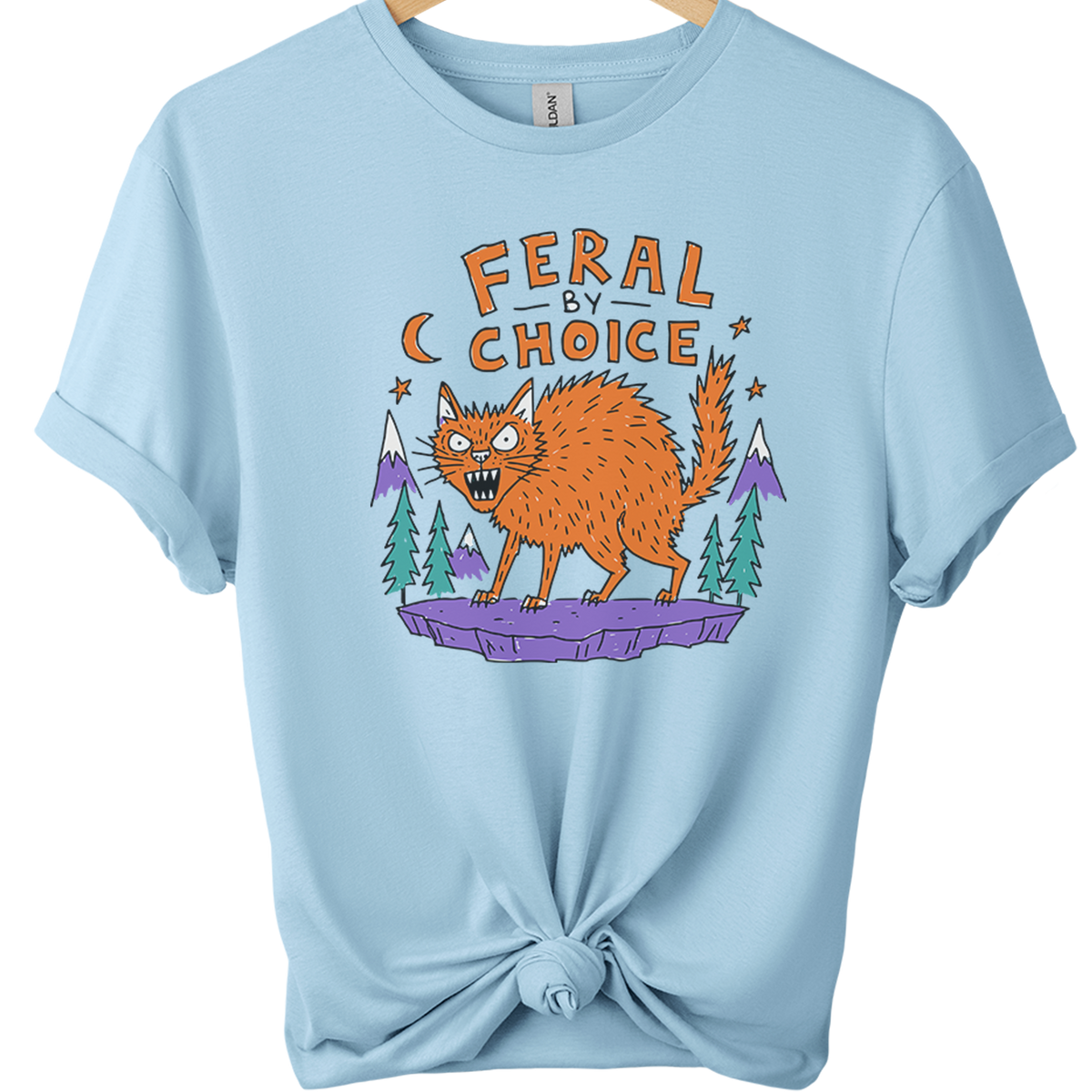 A light blue short-sleeve t-shirt on a wooden hanger, styled with a knot at the bottom hem. The shirt features a graphic print of an orange cat on a purple rock with pine trees and mountains, under the text 'FERAL BY CHOICE'.neilikha
