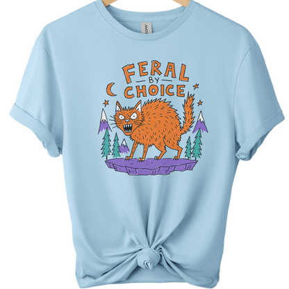 A light blue short-sleeve t-shirt on a wooden hanger, styled with a knot at the bottom hem. The shirt features a graphic print of an orange cat on a purple rock with pine trees and mountains, under the text 'FERAL BY CHOICE'.neilikha
