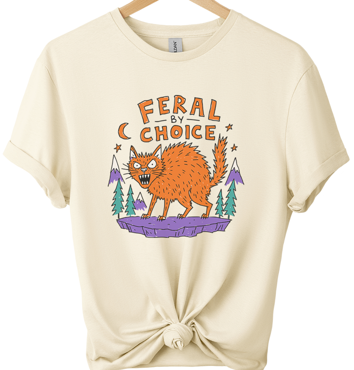 Natural-colored graphic t-shirt on a wooden hanger, styled with a knot at the bottom hem. The shirt features a graphic of an orange cat on a purple rock with pine trees and the text 'FERAL BY CHOICE'.neilikha