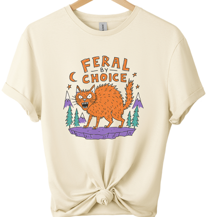 Natural-colored graphic t-shirt on a wooden hanger, styled with a knot at the bottom hem. The shirt features a graphic of an orange cat on a purple rock with pine trees and the text 'FERAL BY CHOICE'.neilikha