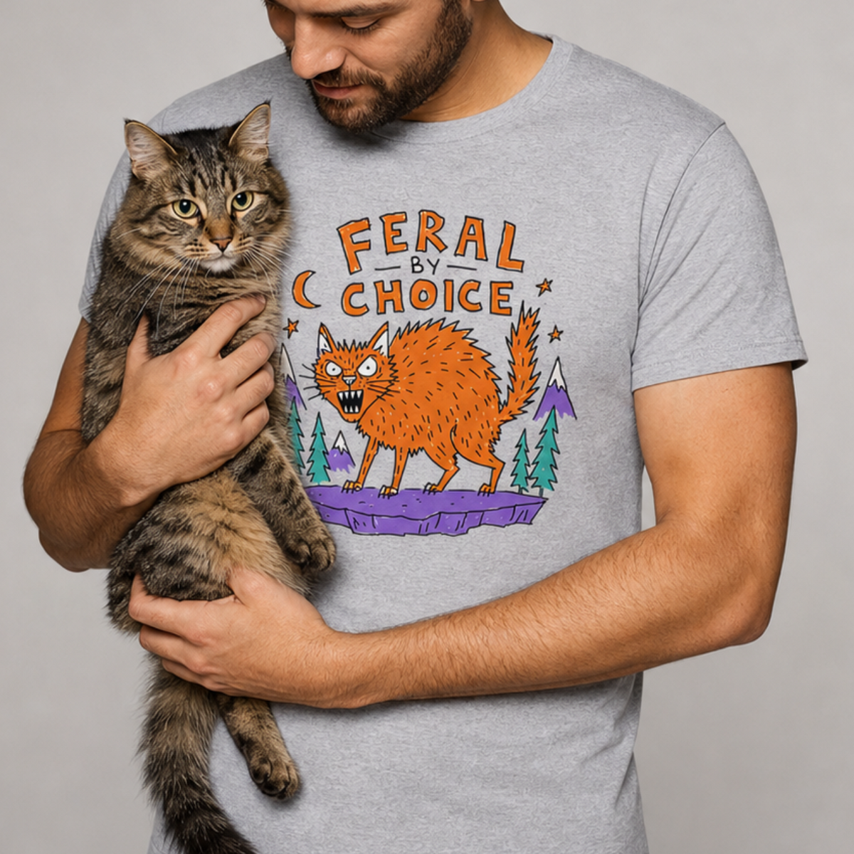 A man wearing a sportgrey graphic t-shirt holds a brown tabby cat against a neutral gray studio background. The t-shirt features a colorful graphic of an orange wild cat with the text 'FERAL CHOICE'.neilikha