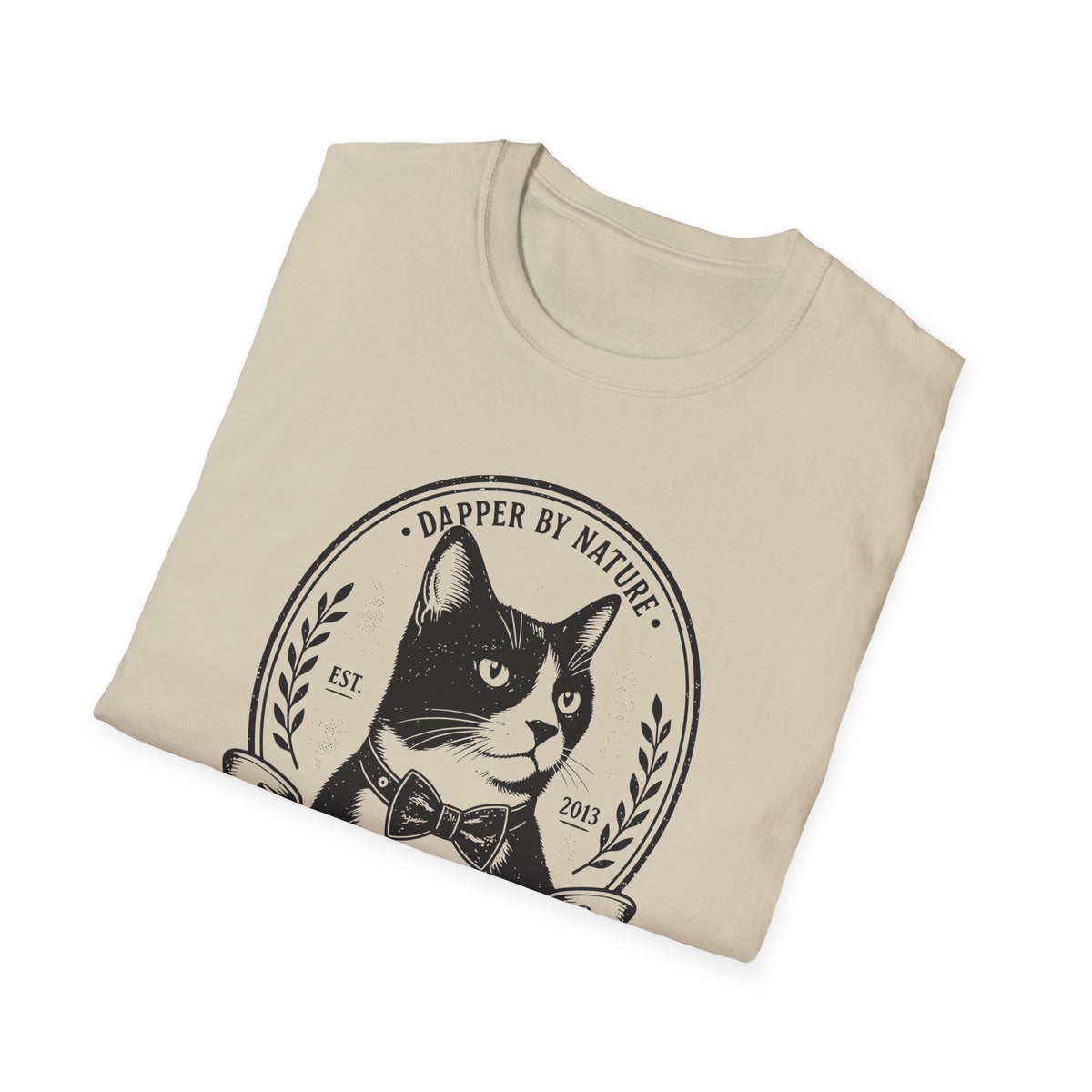 A sand short-sleeve t-shirt folded neatly, featuring a black graphic print of a cat wearing a bow tie inside a circular emblem with the text 'DAPPER BY NATURE'.