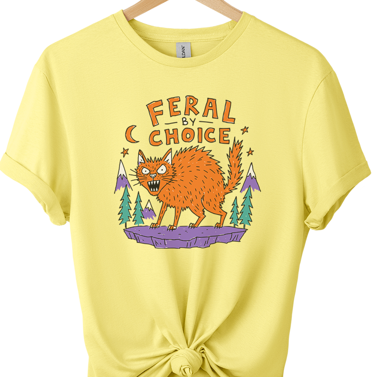 A cornsilk graphic t-shirt on a wooden hanger, styled with a knot at the bottom hem. The shirt features a graphic of an orange werewolf under the text 'FERAL BY CHOICE' with purple mountains and green trees.