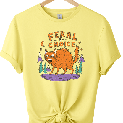 A cornsilk graphic t-shirt on a wooden hanger, styled with a knot at the bottom hem. The shirt features a graphic of an orange werewolf under the text 'FERAL BY CHOICE' with purple mountains and green trees.