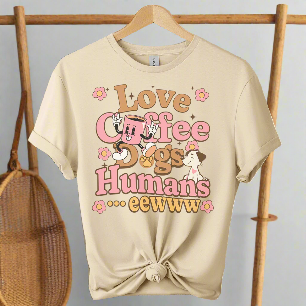 Sand color t-shirt with 'Love Coffee Dogs Humans eewww' text and cartoon graphics on a hanger.