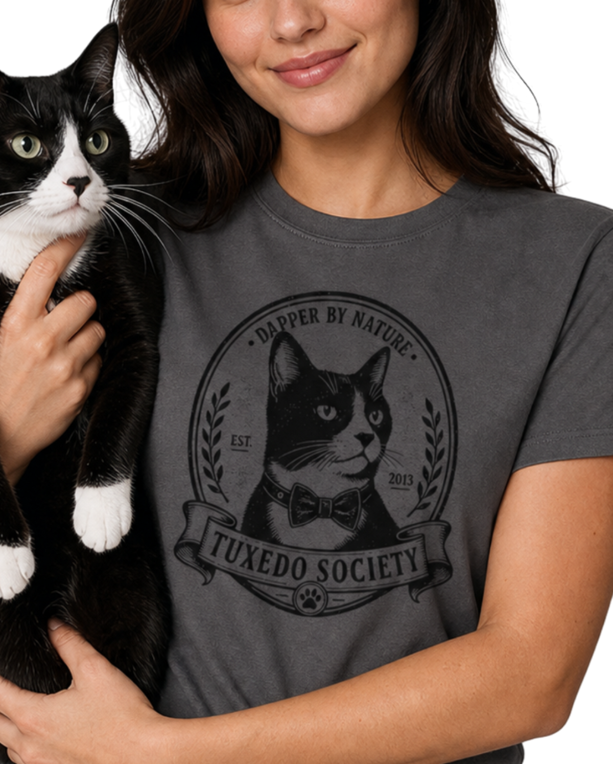 A woman holding a black and white cat wearing a charcoal graphic t-shirt with a 'Tuxedo Society' cat logo. 