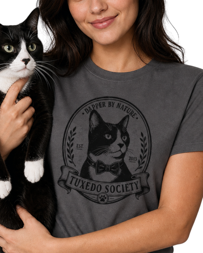 A woman holding a black and white cat wearing a charcoal graphic t-shirt with a 'Tuxedo Society' cat logo. 