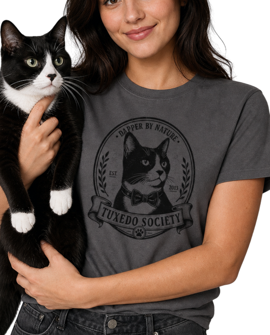 A woman holding a black and white cat wearing a charcoal graphic t-shirt with a 'Tuxedo Society' cat logo. The background is solid white.