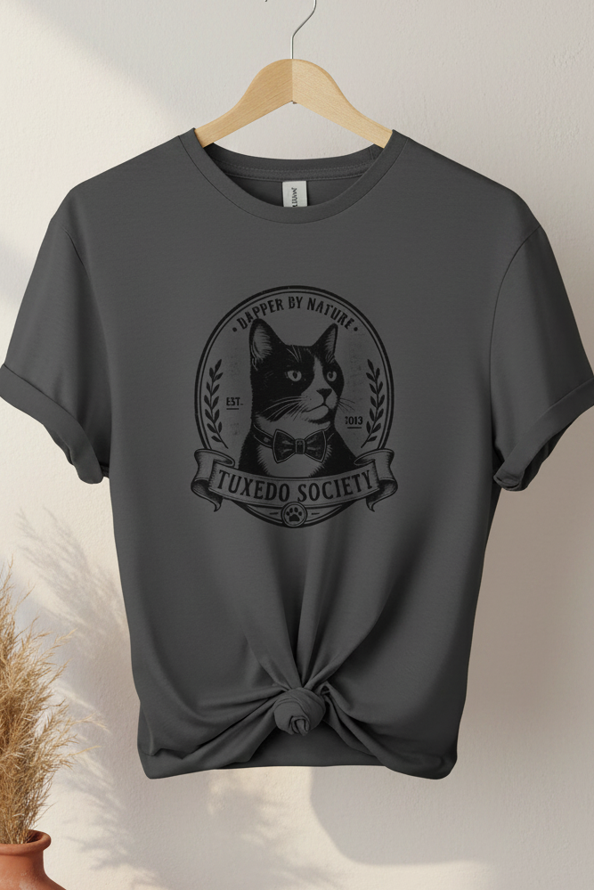 A charcoal t-shirt on a wooden hanger, styled with a knot at the bottom hem. The shirt features a black graphic print of a cat wearing a bow tie inside a circular emblem with the text 'TUXEDO SOCIETY' and 'DAPER BY NATURE'.