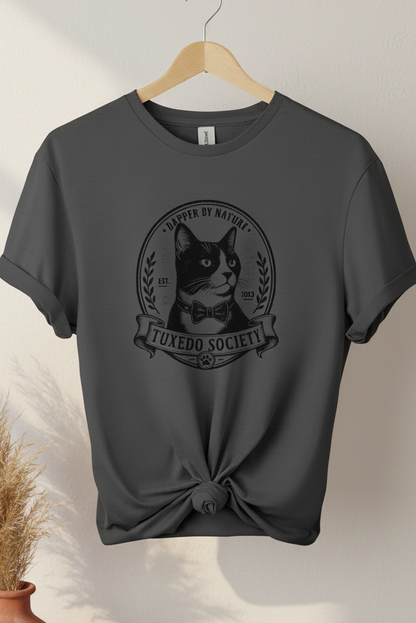 A charcoal t-shirt on a wooden hanger, styled with a knot at the bottom hem. The shirt features a black graphic print of a cat wearing a bow tie inside a circular emblem with the text 'TUXEDO SOCIETY' and 'DAPER BY NATURE'.
