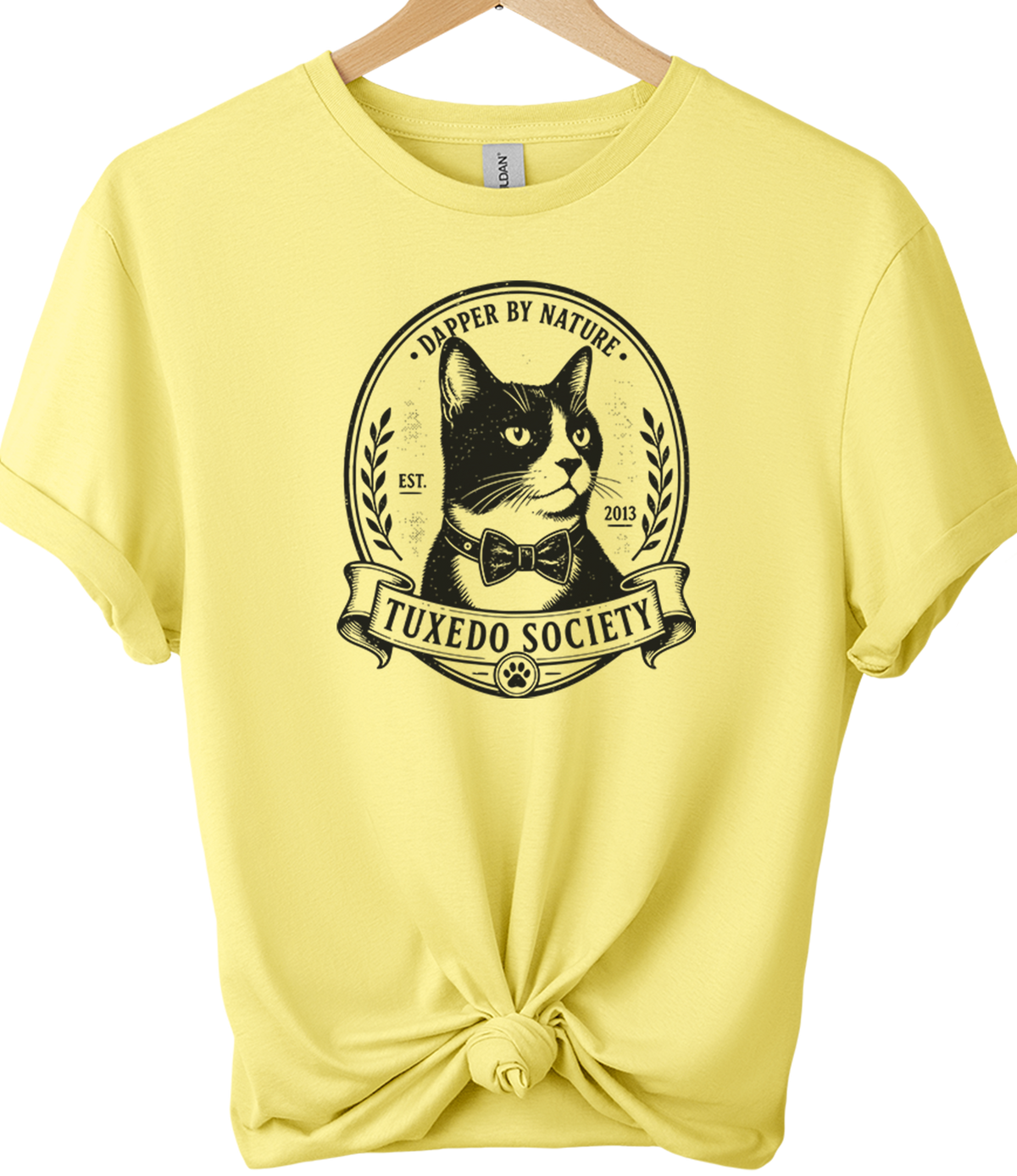 A cornsilk short-sleeve t-shirt on a wooden hanger, styled with a knot at the bottom hem. The front features a black graphic of a tuxedo cat inside an oval frame with the text 'TUXEDO SOCIETY' and 'DAMPER BY NATURE'.