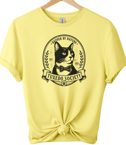 A cornsilk short-sleeve t-shirt on a wooden hanger, styled with a knot at the bottom hem. The front features a black graphic of a tuxedo cat inside an oval frame with the text 'TUXEDO SOCIETY' and 'DAMPER BY NATURE'.